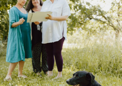 Im Vordergrund ein schwarzer Hund der in der Wiese liegt, hinten drei Frauen die etwas in der Hand halten und sich unterhalten.