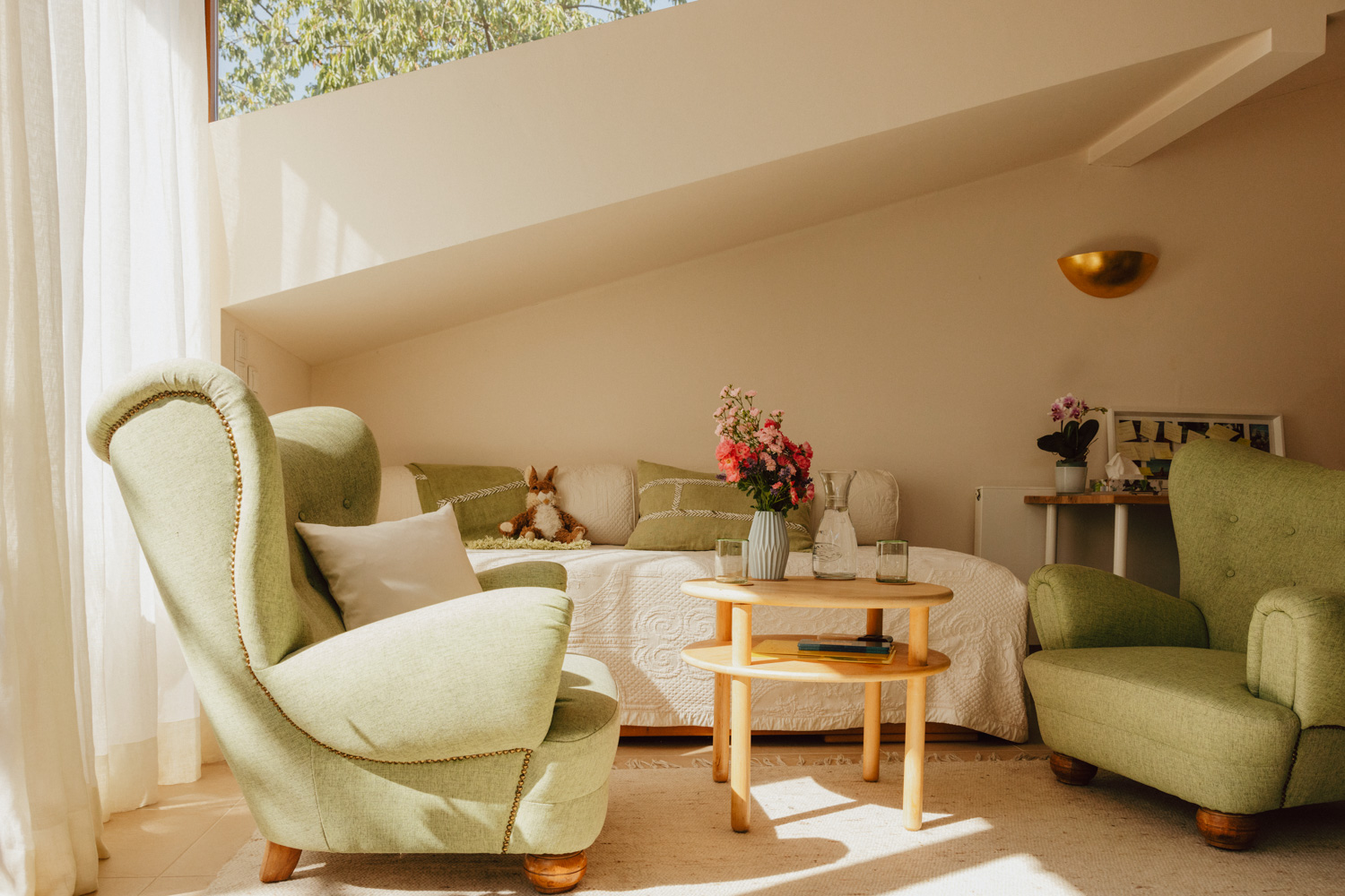 Aufnahme eines Raumes mit großen grünen Ohrensesseln, einer weißen Couch auf dem ein Kuschelhase sitzt und einem runden Holztisch mit frischen rosaroten Blumen.