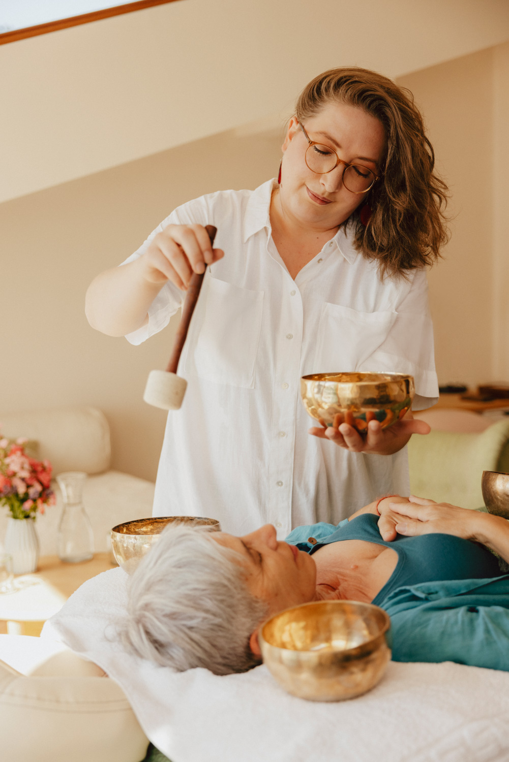 Klangschalen-Klangschalenbehandlung-Klangschalenmassage-Klangbad-Elisabeth-Vogl Foto von mir mit offenen Haaren, weißer Bluse und einem Klöppel in der rechten Hand mit dem ich eine Klangschale in meiner anderen Hand anschlage. Unter der Klangschale liegt eine Frau mit grauen Haaren und einem türkisen Gewand auf einer Massageliege, mit weiteren Schalen die um sie herum aufgestellt sind.