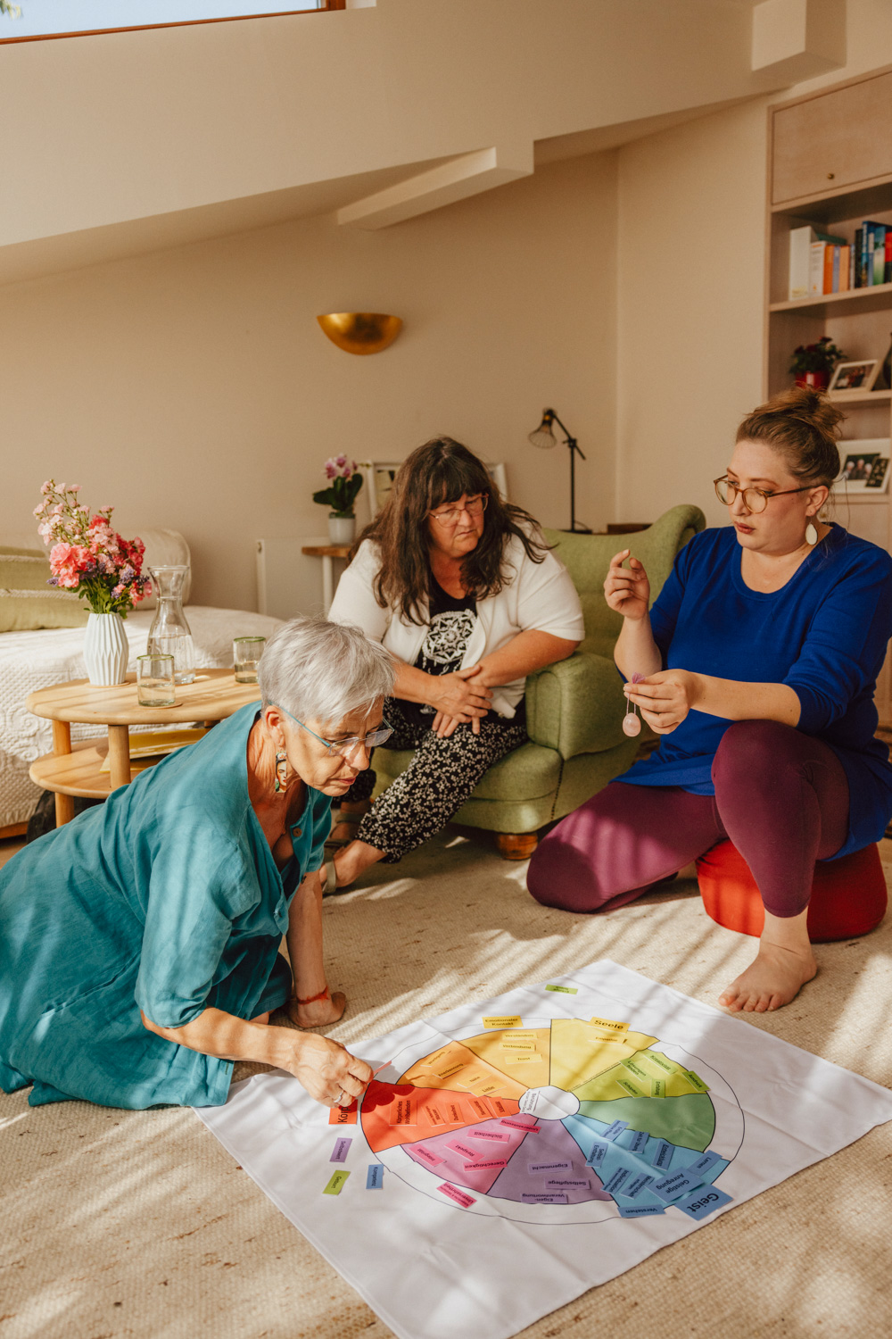 Drei Frauen sitzen und knien um ein buntes Tuch und unterhalten sich. Af dem Tuch sind verschiedene Kärtchen ausgebreitet auf denen Begriffe stehen.