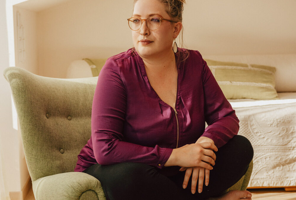 Foto von mir, Elisabeth Vogl, auf einem grünen Ohrensessel sitzend, im Schneidersitz. Mit lila Bluse, schwarzer Leggins und den Blick in die Ferne gerichtet, sieht nachdenklich aus.